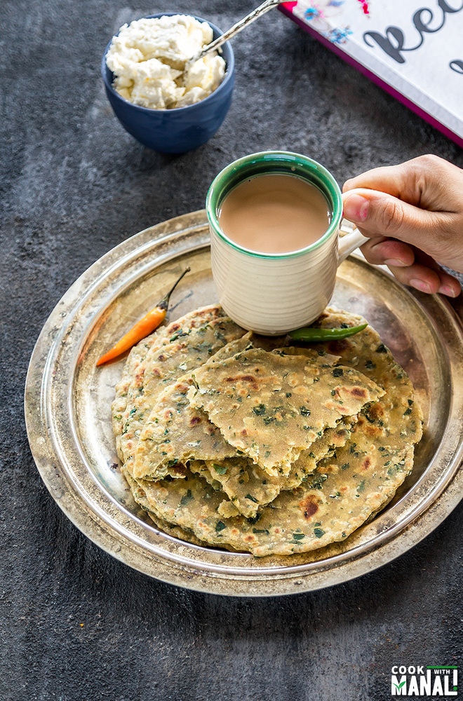 a plate with methi parathas placed on a plate with a hand holding a cup of chai placed on the same plate