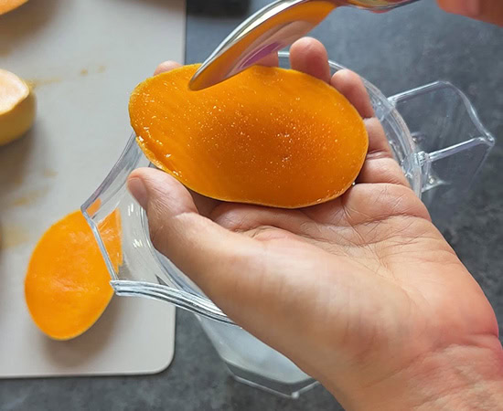 a hand holding a slice of fresh mango and using a spoon to scoop out the mango pulp