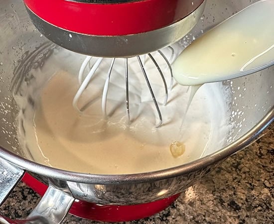 condensed milk being added with a spoon to whipping cream being whipped in mixer
