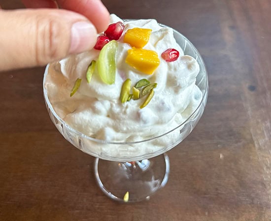 a hand adding fruits and nuts on top of whipped cream served in a glass bowl